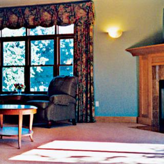 A cozy living room with an armchair, a wooden fireplace, and large windows adorned with floral curtains.
