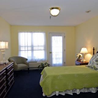 A cozy bedroom with a bed, dresser, chair, and lamps in a softly lit space.