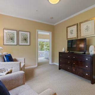 A cozy living room with a beige color scheme, featuring a sofa, wall art, a television on a dark wooden dresser, and carpeted flooring.