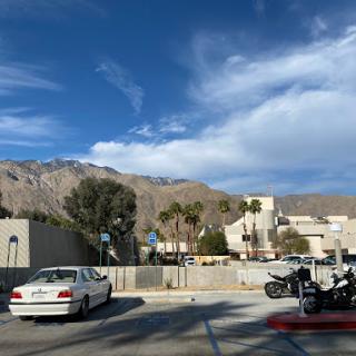 A serene view of mountains under a partly cloudy sky, featuring vehicles parked in a lot and palm trees in the foreground.