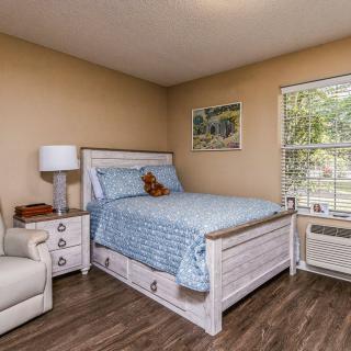 A cozy bedroom featuring a bed with blue bedding, a nightstand with a lamp, and a comfortable chair by the window.