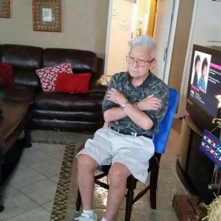 An elderly person sitting in a living room with crossed arms, watching television.