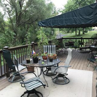 A cozy outdoor deck area with a table, chairs, and an awning surrounded by lush greenery.
