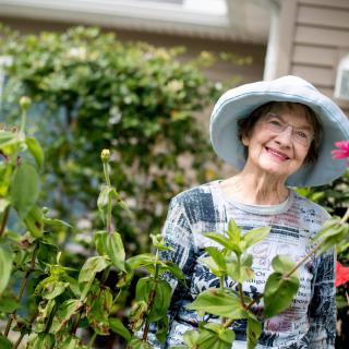 An elderly woman wearing a hat smiles while standing in her garden surrounded by lush greenery and flowers.