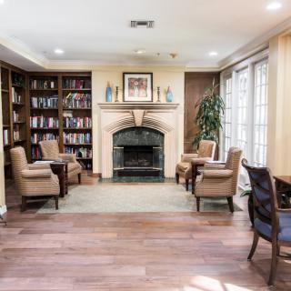 A cozy library setting with a fireplace, surrounded by bookshelves and comfortable lounge chairs, illuminated by natural light from large windows.