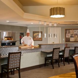 A chef stands behind the counter in a restaurant kitchen with dining area seating.