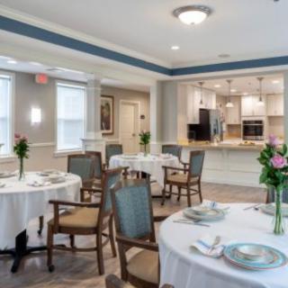 An elegantly set dining area with neatly arranged tables and chairs, featuring floral centerpieces and a view of the kitchen.