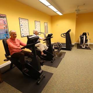 Senior citizens exercising on recumbent bikes in a gym with yellow walls.