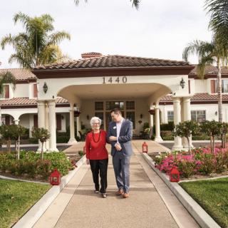 A senior woman and a man walking together on a pathway in front of a residential building with well-maintained landscaping.