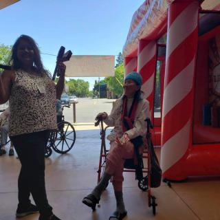 A person with a microphone stands next to someone sitting in a wheelchair near a bouncy castle outdoors.
