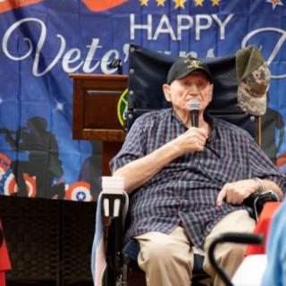 An elderly veteran speaks at a Veterans Day celebration, holding a microphone in front of an American flag backdrop.