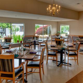 A spacious restaurant dining area with neatly arranged tables and chairs, illuminated by a chandelier and natural light from large windows.