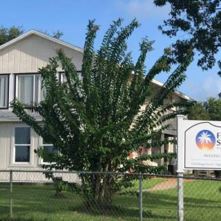 A two-story building with a large tree and a sign in the front yard, surrounded by a chain-link fence.