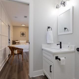 A modern, minimalist bathroom with a white vanity and wood flooring opens into a cozy room.