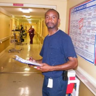A healthcare worker in scrubs holds a clipboard in a hospital hallway.
