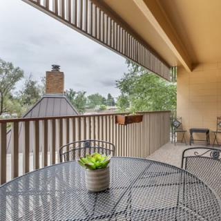 A cozy balcony with a metal table and chairs, featuring a potted plant and overlooking a view with trees under an overcast sky.