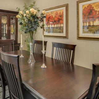 A well-decorated dining room featuring a dark wood table with matching chairs, adorned with a vase of flowers and candles. The walls are decorated with colorful landscape paintings.