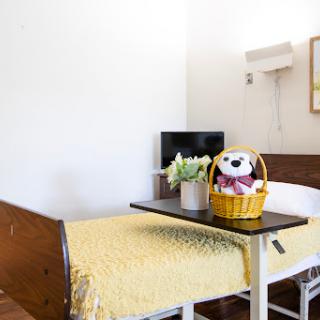 A cozy hospital room featuring a patient bed adorned with a yellow blanket, a small table with a decorative basket and stuffed animal, and a TV in the background.