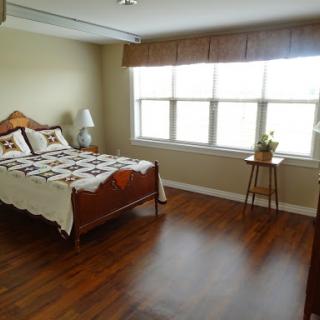A cozy bedroom featuring a neatly made bed, warm wooden flooring, and natural light coming through large windows.
