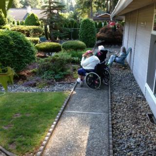 Two people sitting outdoors near a garden and a house, one in a wheelchair on a pathway surrounded by greenery.
