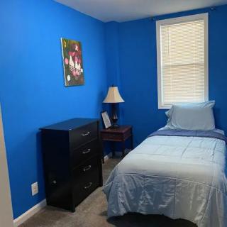 A small bedroom featuring blue walls, a single bed, a lamp on a side table, and a chest of drawers.