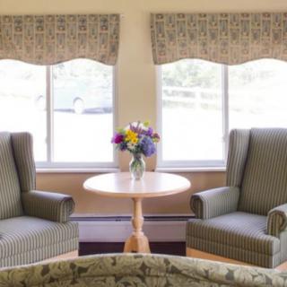 A cozy living room setup with two armchairs and a small round table adorned with a vase of colorful flowers in front of large windows.