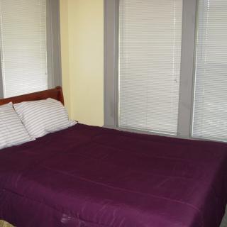 A neatly made bed with purple bedding and pillows beside windows with closed blinds.