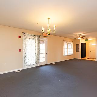 An empty room with carpet flooring, featuring a chandelier and ceiling fan, with windows and a doorway.