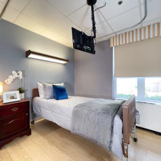 A well-lit hospital room featuring a single bed, wooden bedside table, and wall-mounted television.