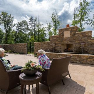 Two people sitting on a patio surrounded by nature, enjoying a peaceful outdoor setting with a large stone fireplace.