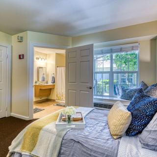A cozy bedroom with a neatly made bed, accented by pillows and a blanket, features a window overlooking greenery. An adjacent bathroom is visible through an open door.