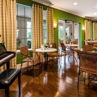 A cozy dining area with a piano, featuring wooden floors and large windows adorned with curtains.