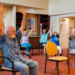 A group of seniors participating in a seated exercise session, raising their arms together in an indoor setting.