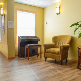 A cozy waiting area featuring a comfortable armchair, piano, side table, and indoor plant under warm natural light.