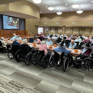 A group of elderly individuals in wheelchairs gathered at tables for a community event in a dining hall.