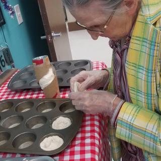 An elderly woman is preparing dough in a muffin tray on a checkered tablecloth.