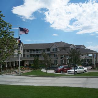 A three-story building with cars parked in front, an American flag flying, and a well-maintained lawn under a partly cloudy sky.