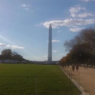 A view of the Washington Monument on a sunny day with clouds in the sky and people walking along a pathway.