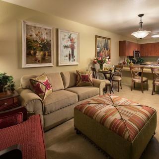 A cozy living room with a comfortable sofa and large ottoman, decorated with artwork on the walls and an open view of a kitchen area.
