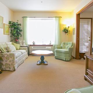 A cozy living room with light green and neutral tones, featuring a sofa, armchair, coffee table, and large window with curtains.