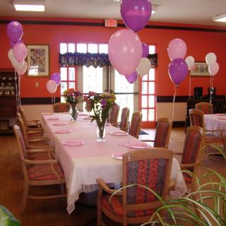 A beautifully decorated dining room set up with pink and purple balloons and floral centerpieces for a celebration.