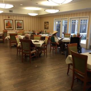 A quiet dining room with tables and chairs, occupied by a few seniors.