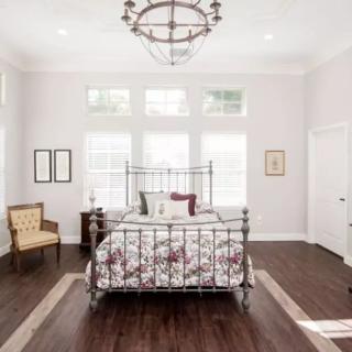 A spacious and elegantly designed bedroom featuring a metal bed frame, floral bedding, and a large chandelier. The room is adorned with wooden flooring and white walls, enhanced by natural light from multiple windows.