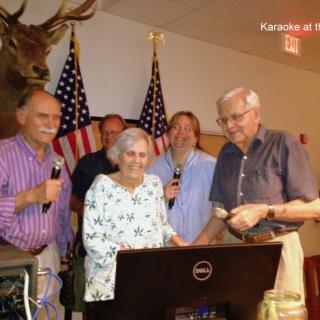 A group of elderly individuals gathered indoors, holding microphones, with American flags in the background.