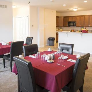 A dining area with tables set for a meal, featuring red tablecloths and leather chairs, adjacent to a kitchen.