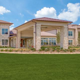 A modern residential building with a large, covered entrance and well-maintained lawn under a blue sky.