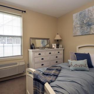 A cozy bedroom with a neatly made bed, a dresser topped with a lamp and mirror, and a window letting in natural light.