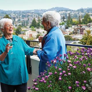 Two elderly women enjoying a conversation and a glass of wine on a balcony overlooking a cityscape.