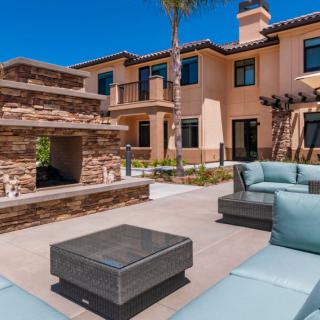 A luxurious outdoor patio with modern furniture and a stone fireplace, set against a stylish two-story house on a sunny day.