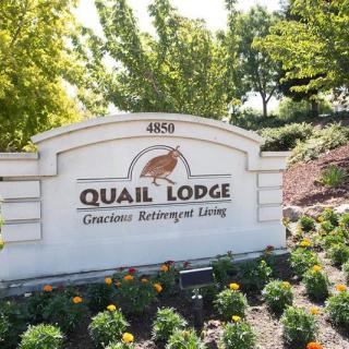 A sign for Quail Lodge Senior Retirement Living surrounded by trees and flowers.